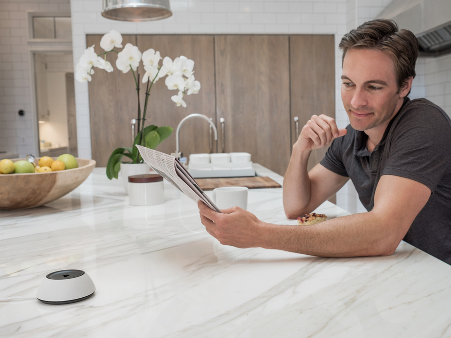 A man sitting in a kitchen reading a newspaper with a Josh.ai device on the counter.